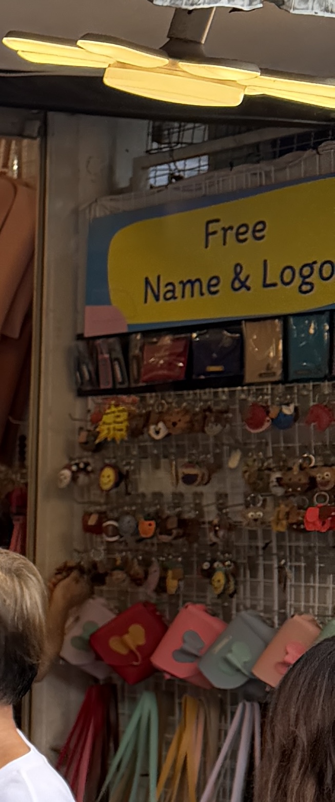 チャトゥチャック市場の革小物店。Free Name and Logoと書かれた黄色い看板の下にキーホルダーが並ぶ