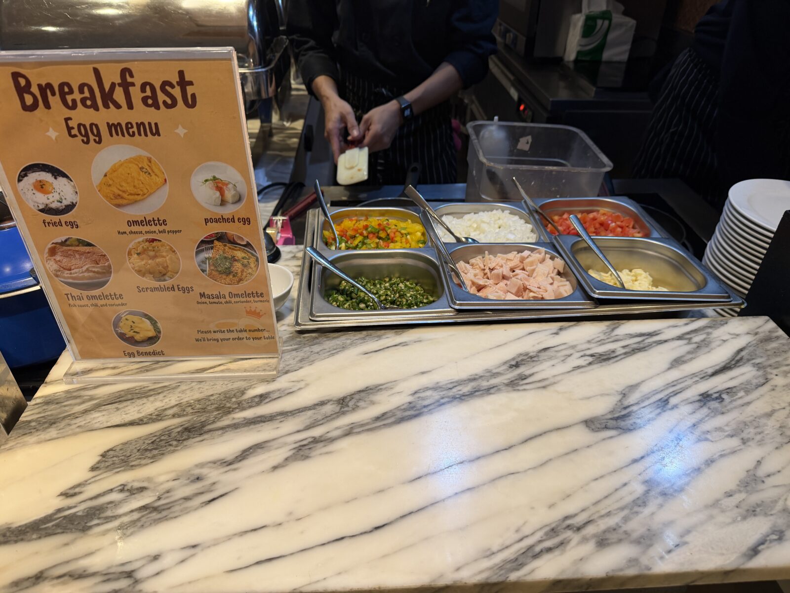 モーベンピックの朝食の卵料理カウンター。Breakfast Egg menuの案内と、具材を入れたバットが並ぶ