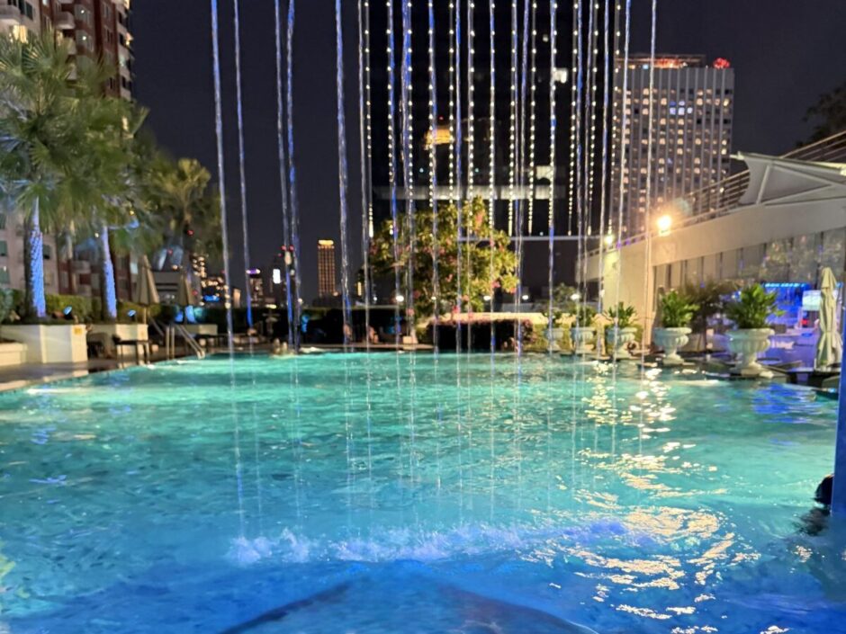 ザ・バークレー・ホテル・プラトゥーナムの夜のプール。上から水のカーテンが落ち、照明で青く光る水面の奥にバンコクのビルの夜景が広がる