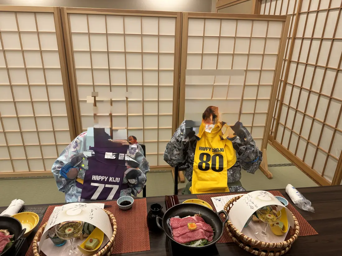 和室の食事席に、喜寿の紫と傘寿の黄色の名入れTシャツが飾られ、前に夕食の膳が並ぶ