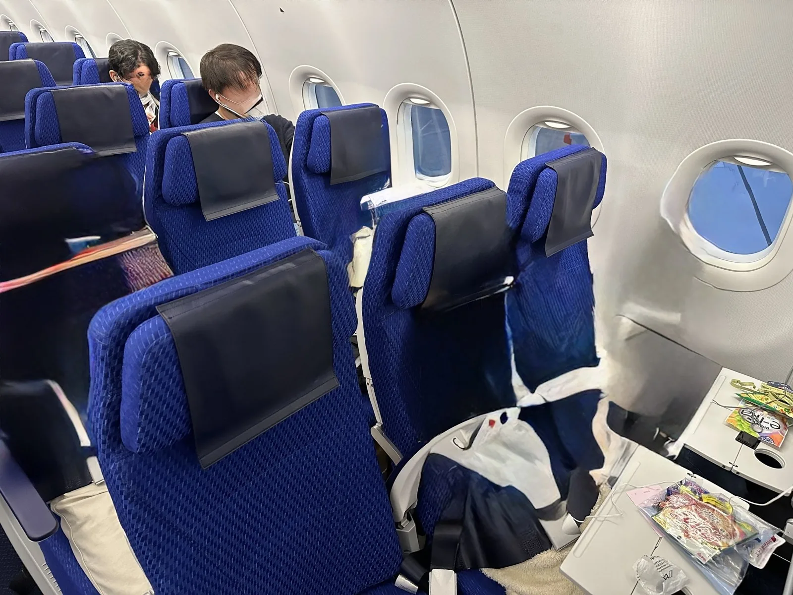 旅客機の客室に並ぶ青い座席と窓。手前の座席テーブルの上に菓子の袋が置かれている