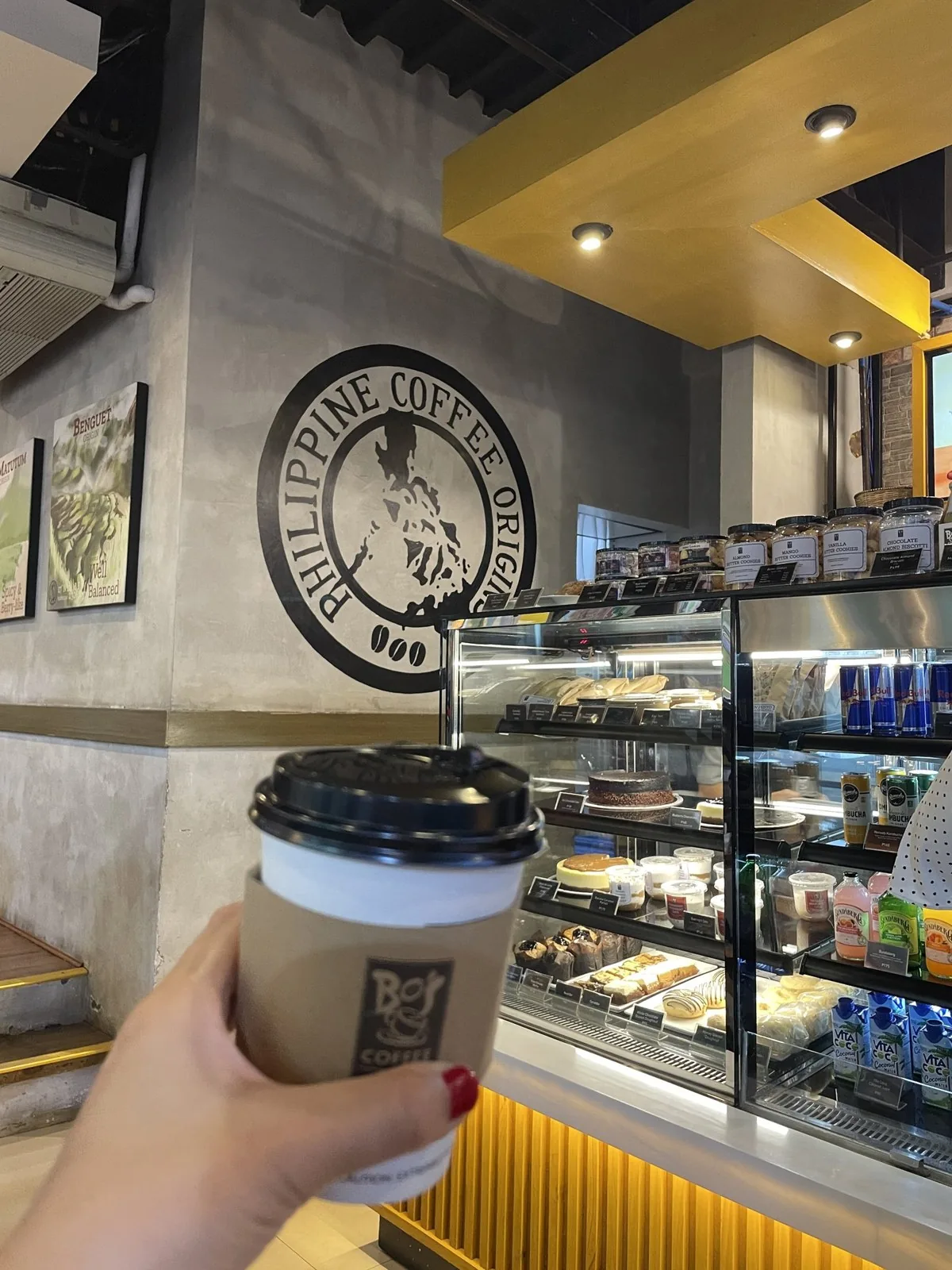 BO'S COFFEE のカップを手に持っている。奥の壁にフィリピンの形のロゴがある