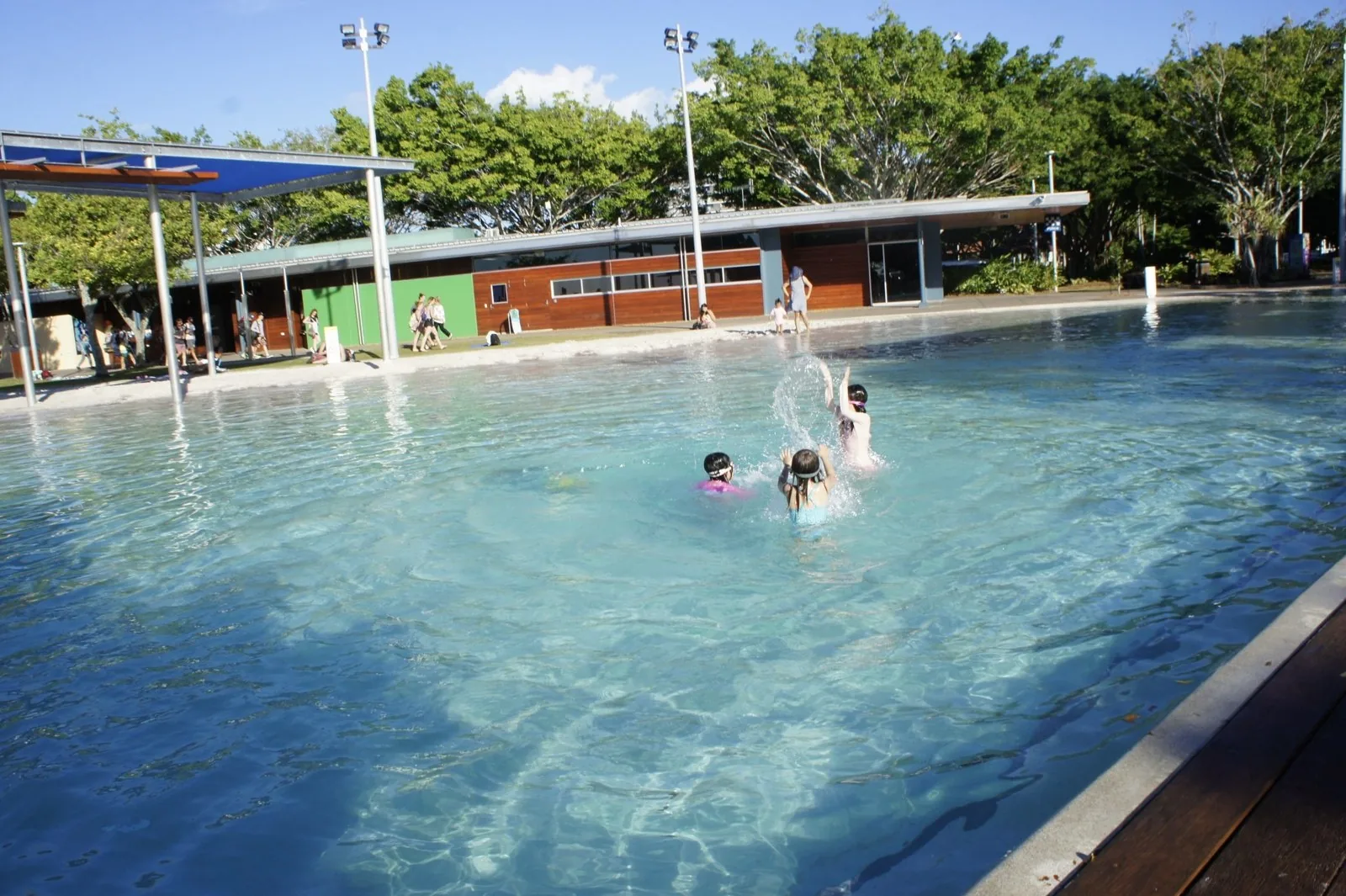 Three children splashing in the outdoor lagoon pool surrounded by white sand, with buildings and trees behind