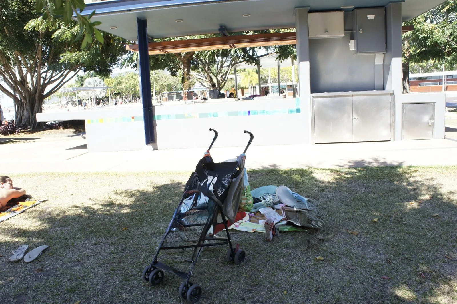 A black stroller on the grass and our things on a picnic mat, with a roofed building and trees behind