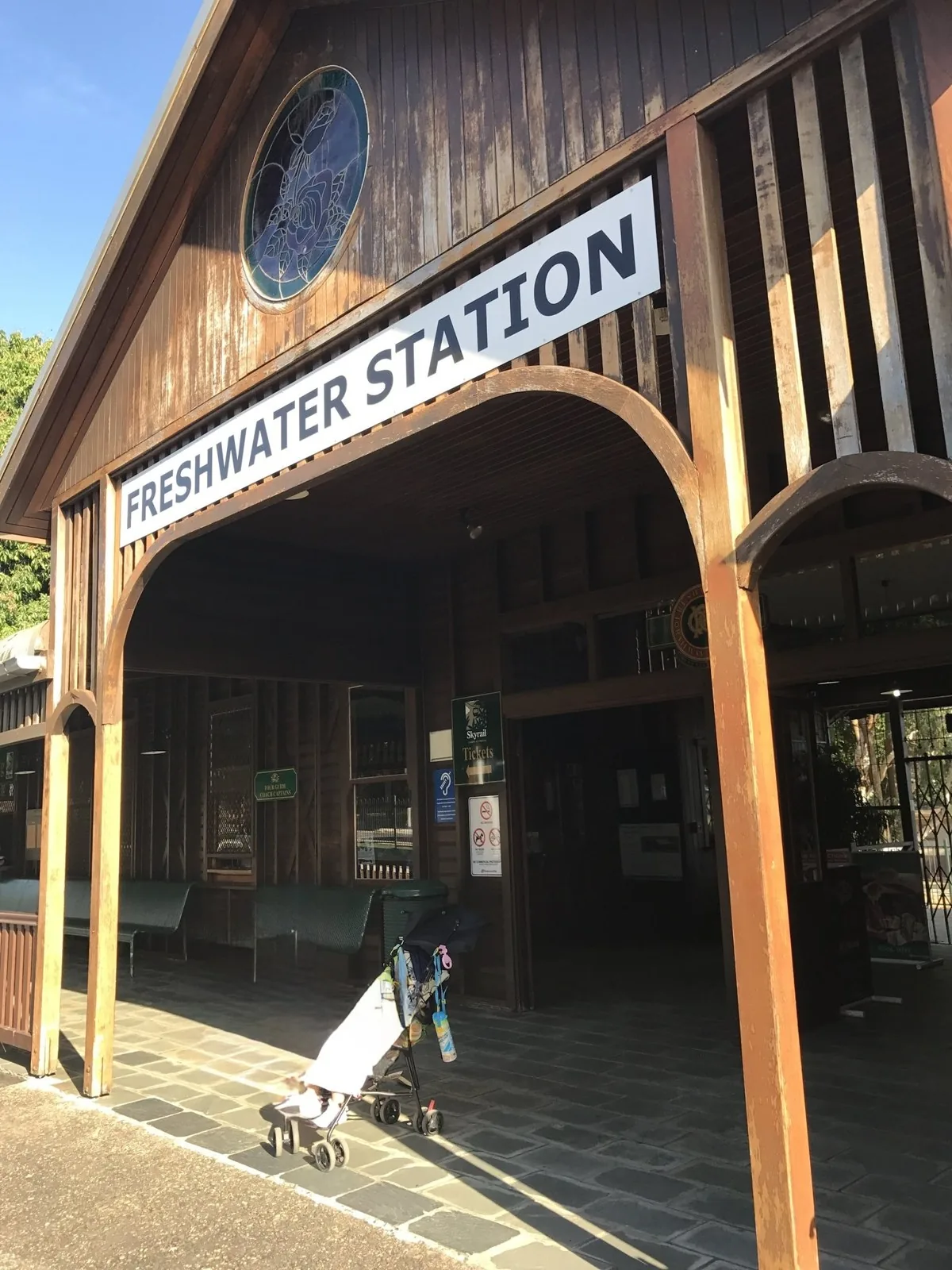 The entrance of a wooden station building with a FRESHWATER STATION sign and a round stained-glass window, with a stroller in front