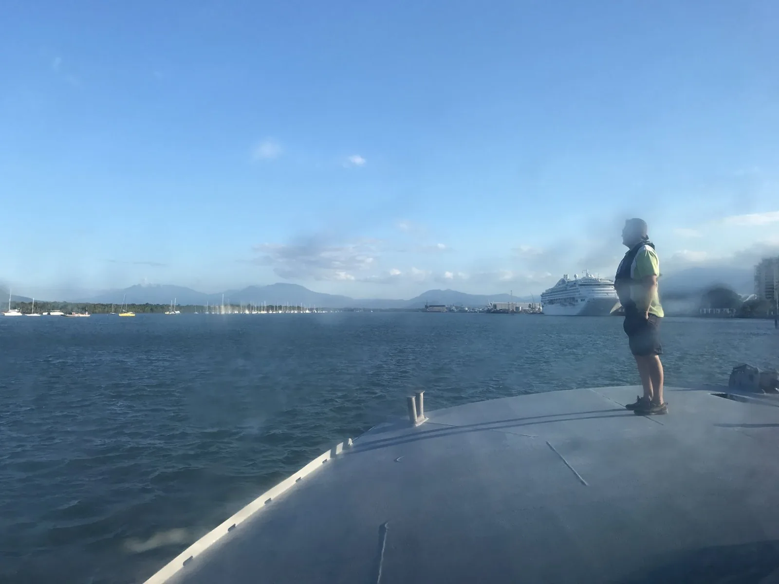 The harbor seen from the boat's deck, with a large cruise ship moored, a shore lined with yachts, and mountains behind