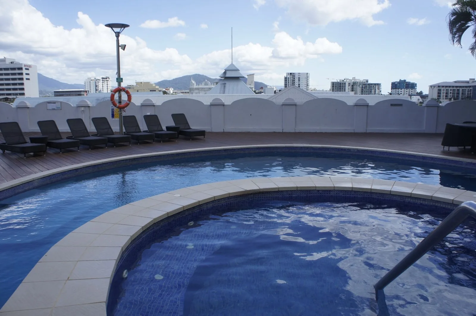 Pool with blue tiles and a jacuzzi, a poolside lined with deck chairs, and the city and mountains beyond