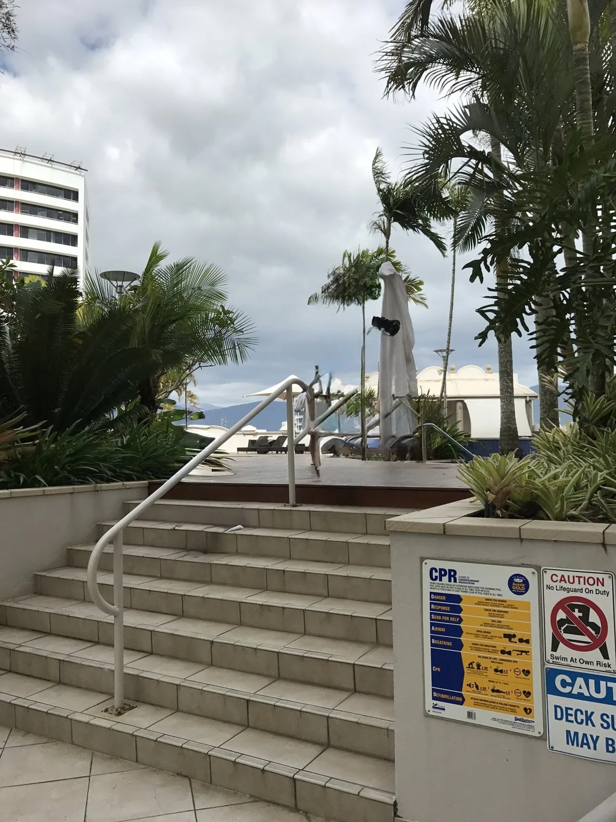 Tiled stairs with white railings leading up to the hotel pool deck, surrounded by palm trees, with a warning sign