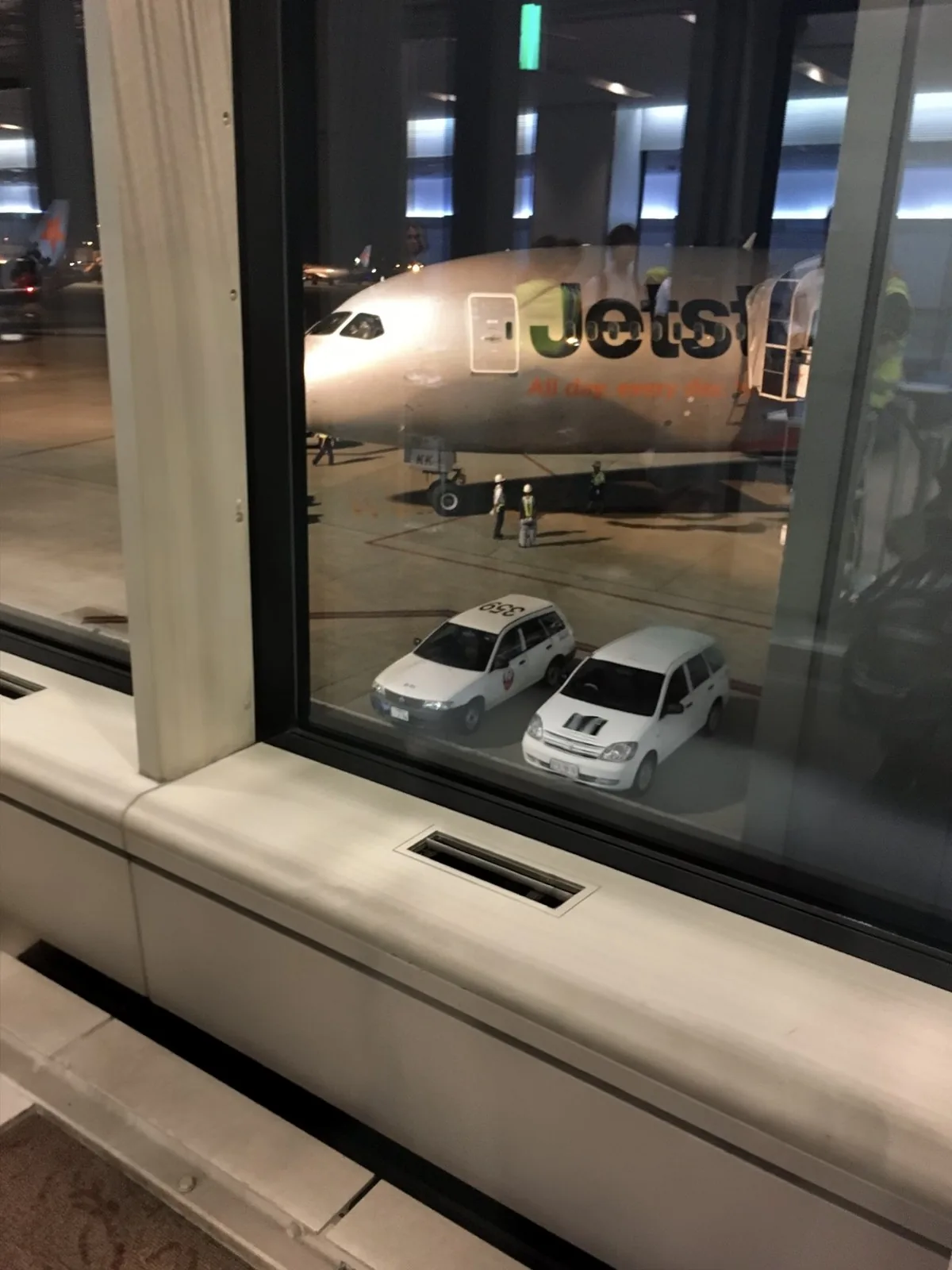 空港ターミナルの窓越しに見える、夜の駐機場のジェットスター機と白い作業車