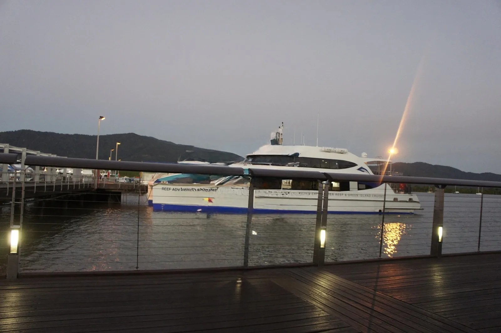 A white catamaran moored at the harbor, seen over the railing of a wooden deck at dusk, with mountains behind