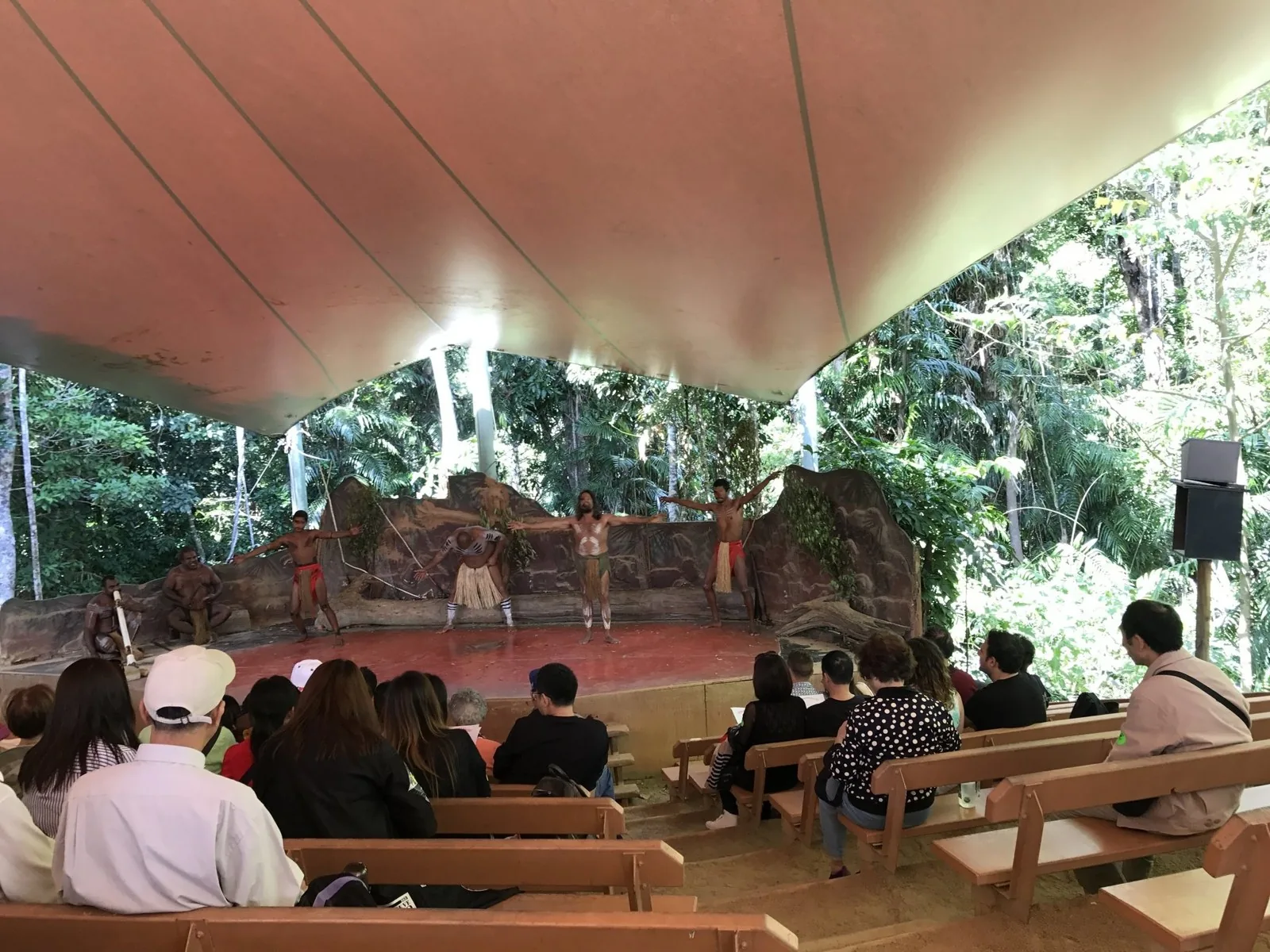 Body-painted performers dancing on a tent-roofed stage in the rainforest, with the audience watching from benches