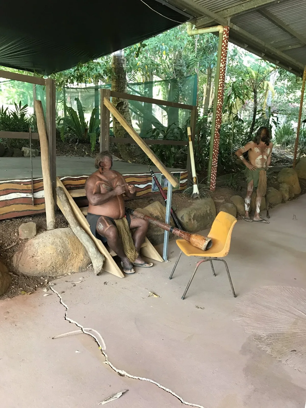Two body-painted men and a didgeridoo leaning against a yellow chair at a covered show venue