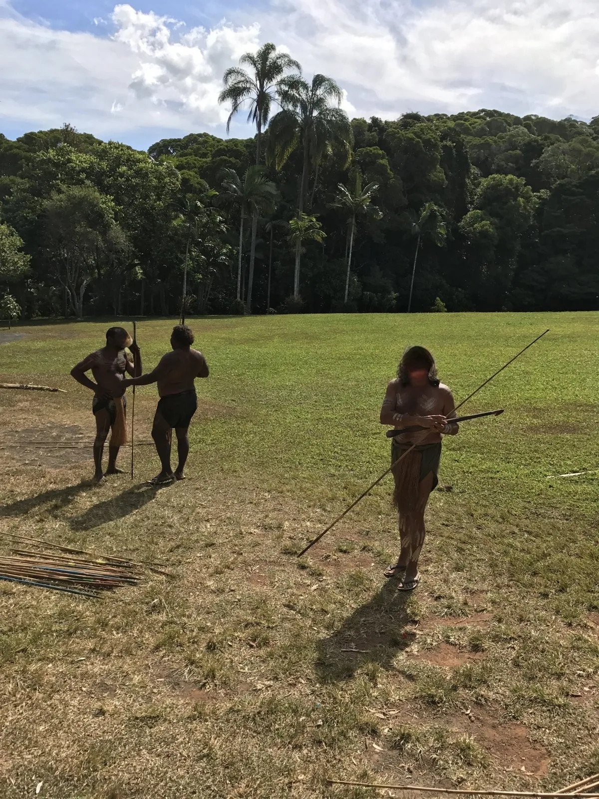 Body-painted men standing with spears and spear throwers on a grassy area with the rainforest behind