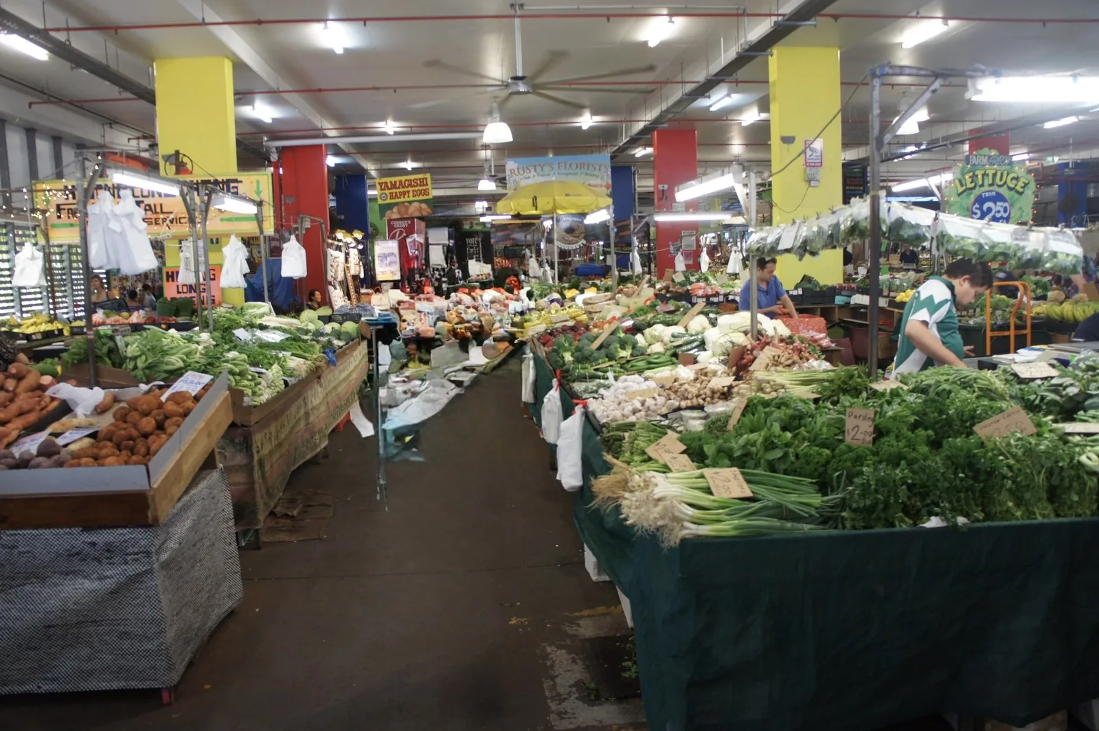 Stalls of vegetables such as spring onions, parsley, and potatoes lining both sides of an aisle in an indoor market