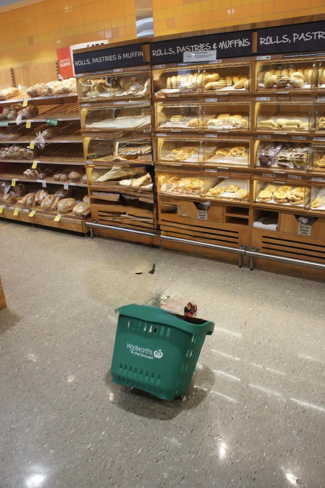 A supermarket bakery section with bread rolls and sweet buns, and a green Woolworths shopping basket on the floor