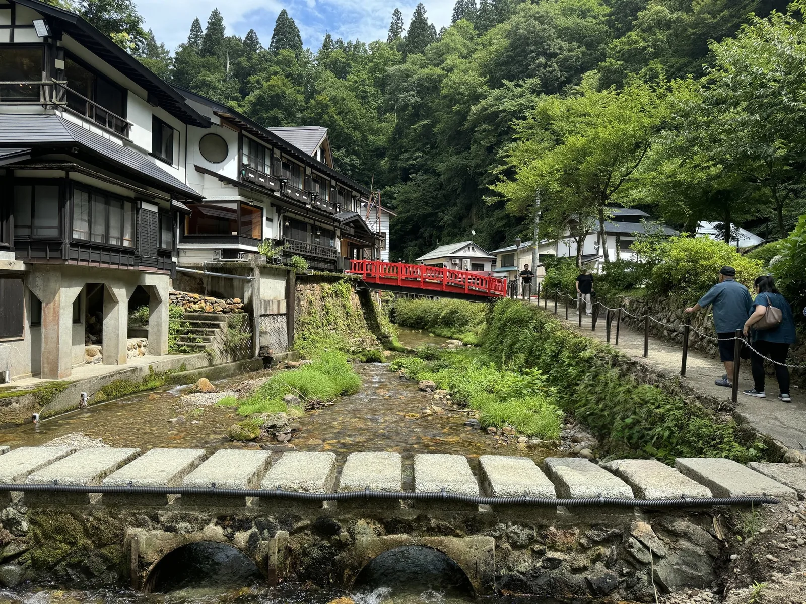 夏の銀山温泉。銀山川に沿って木造の旅館が並び、奥に赤い欄干の橋、手前に石の橋が見える
