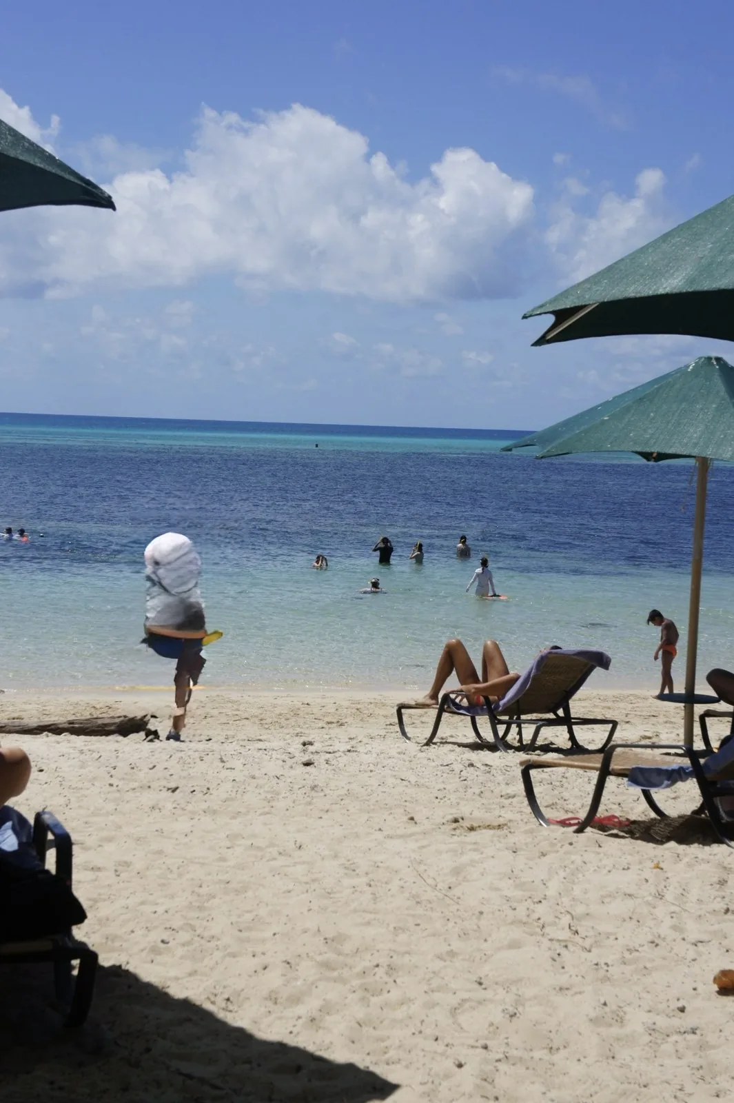 A white sand beach lined with green umbrellas and beach chairs, and clear blue water with people snorkeling