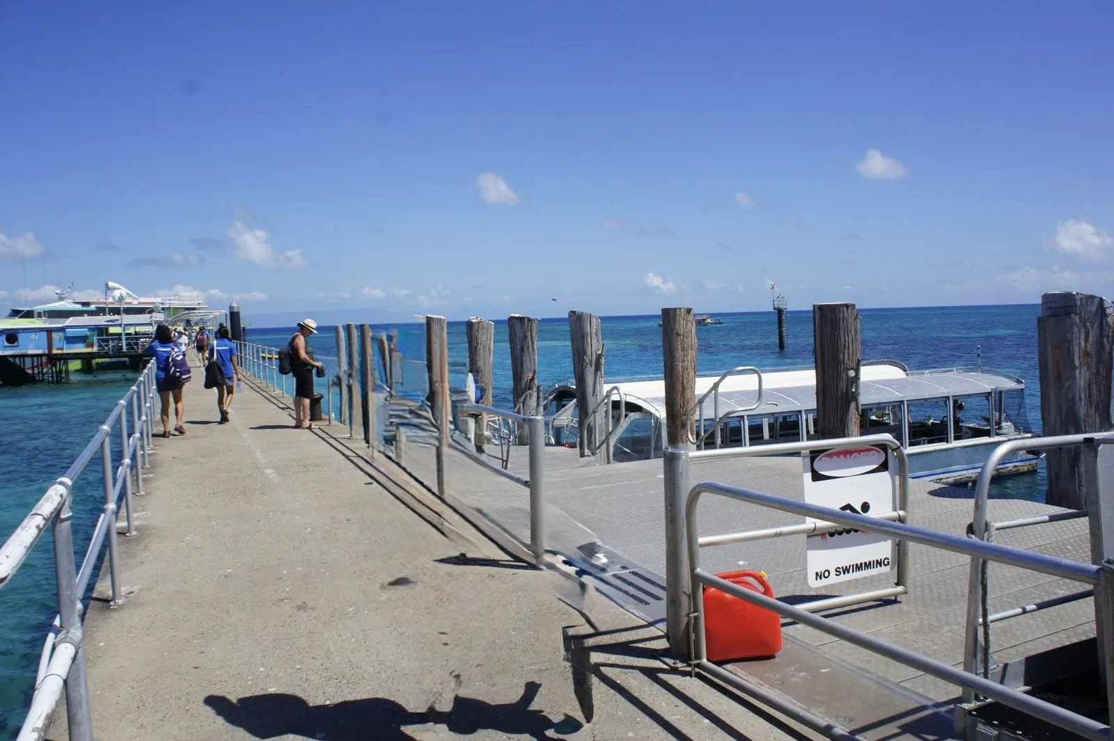 A jetty running out into turquoise water, a white-roofed boat tied up alongside, and a large ferry moored behind