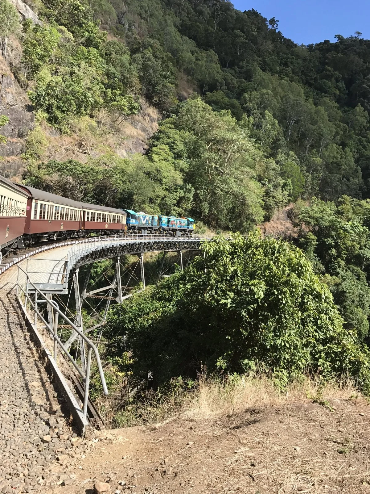 熱帯雨林の山肌に沿ってカーブする鉄橋を渡る、青い機関車と茶色の客車の列車