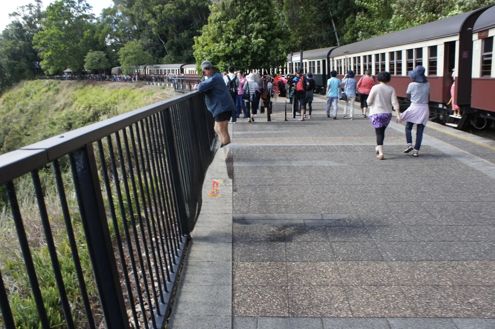 A platform along the tracks with maroon and cream carriages stopped, and tourists walking beside the fence