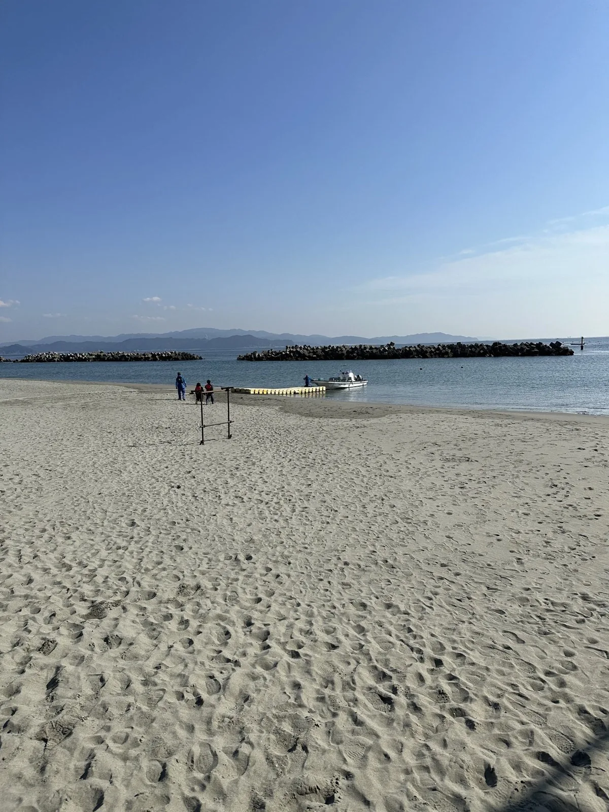 足跡の残る砂浜の先に、消波ブロックの防波堤と黄色い浮桟橋、小型ボートが見える青空の海辺