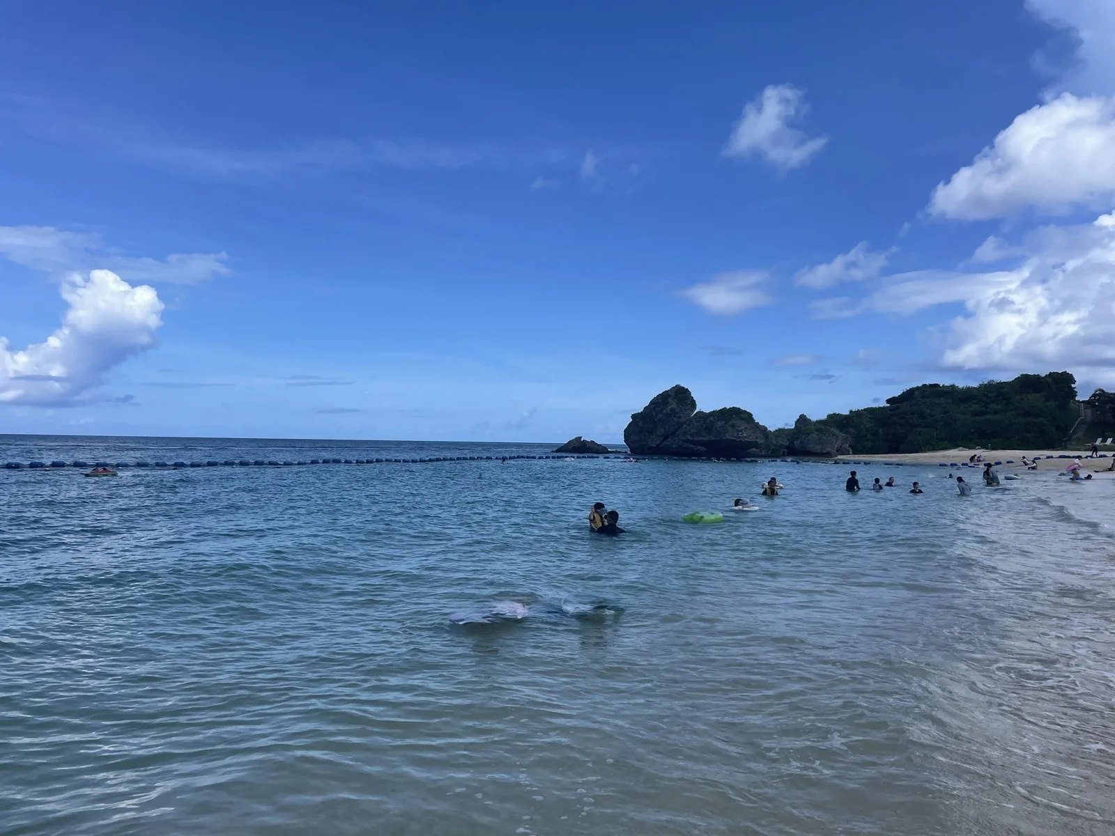 ニライビーチの、ブイで囲まれた遊泳区域の青い海と奥の岩場、浅瀬で泳ぐ人たちと晴れた青空