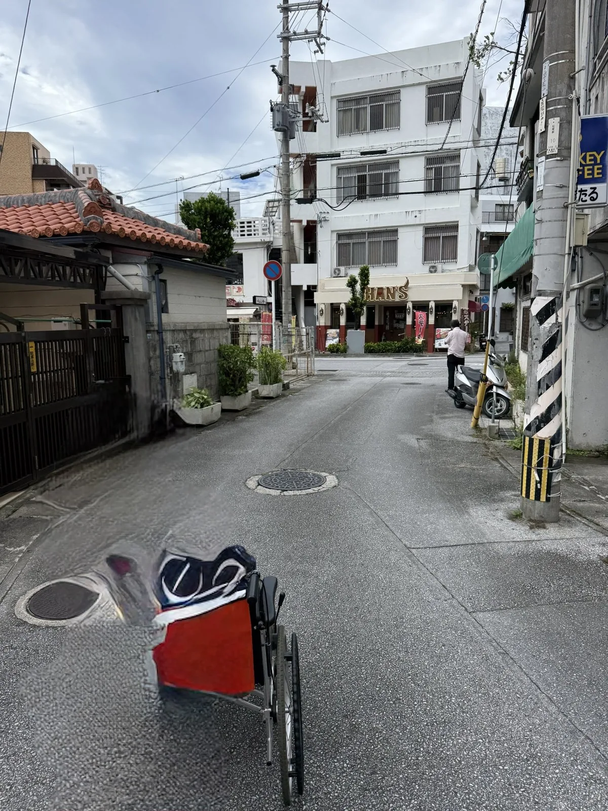 赤瓦屋根の家と白いビルに挟まれた路地と、手前に置かれた荷物を載せた車椅子