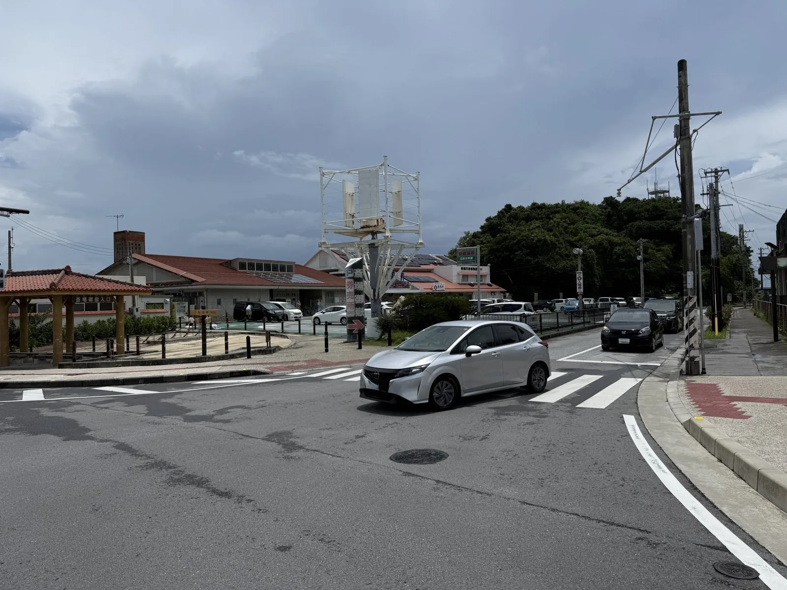 斎場御嶽入口の看板が掛かった東屋と、南城市地域物産館前の交差点を走る銀色の車