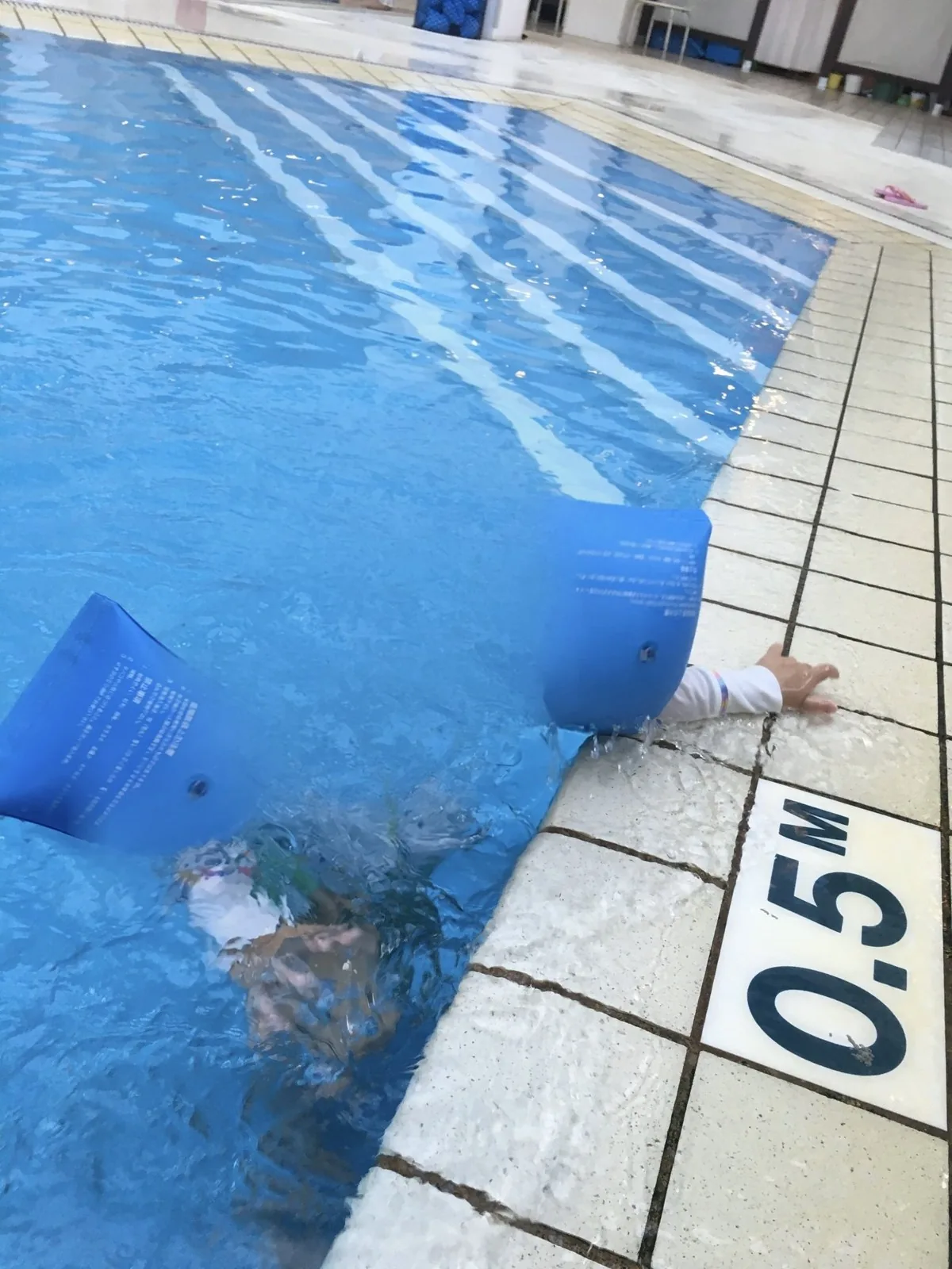 水深0.5mの表示があるプールの縁と、水につかる小さな子