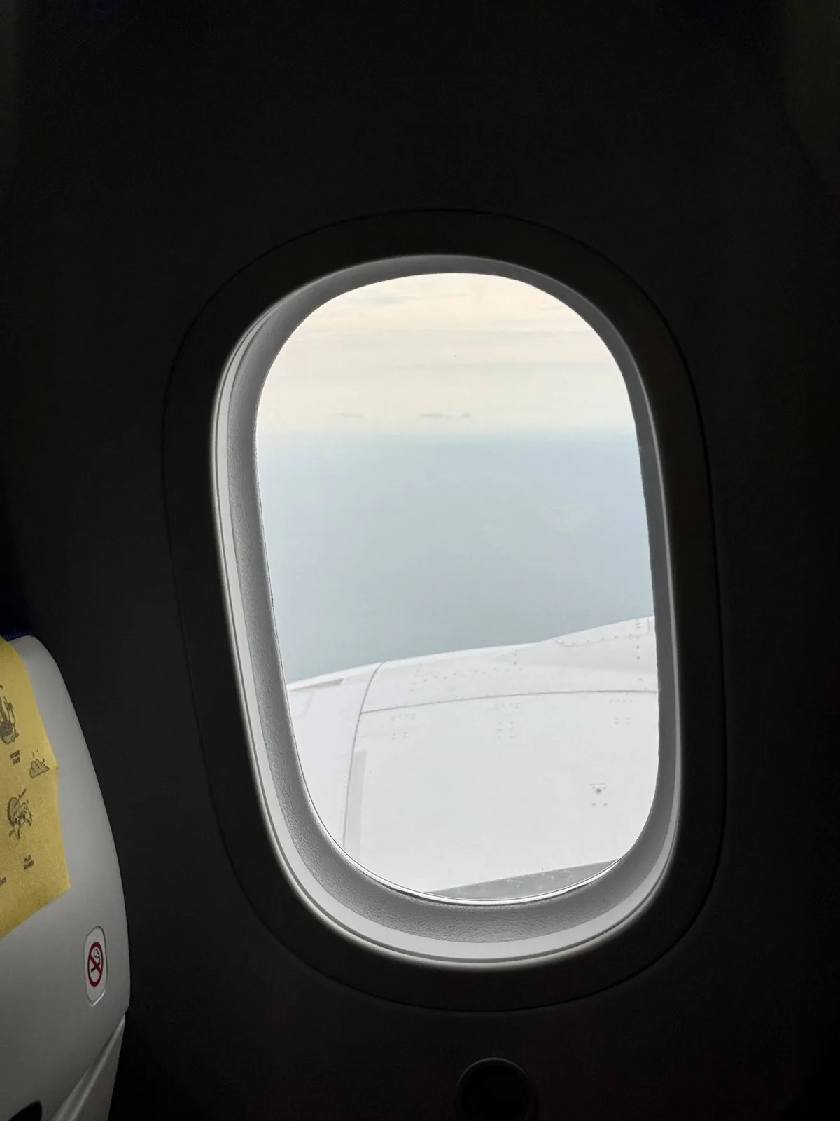 A white wing seen from an airplane window, with a hazy sea and a cloudy sky beyond it