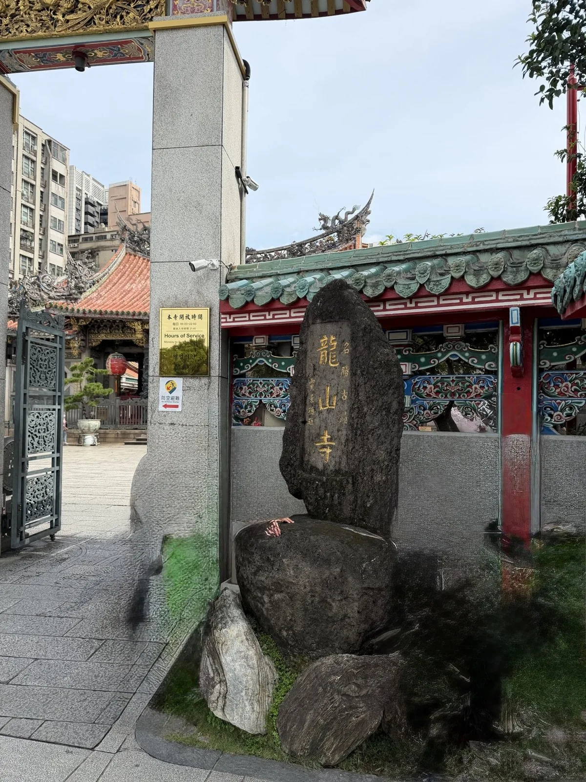 A stone monument carved with the temple name in gold letters, with the red-tiled temple buildings visible beyond the gate