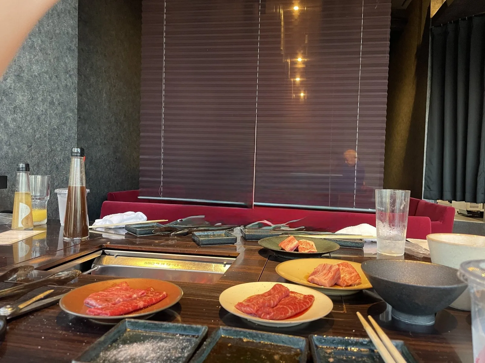焼肉店のロースター付きのテーブルに並ぶ霜降りの牛肉の皿とタレの小皿、奥に赤いソファが見える